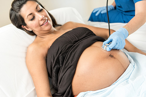 A pregnant patient lying down as a health care provider listens to her stomach using a stethoscope.