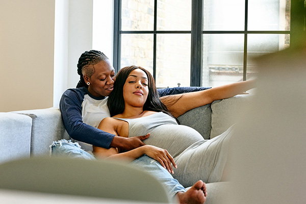 A pregnant woman lying down on a couch in the arms of a woman also sitting on the couch.