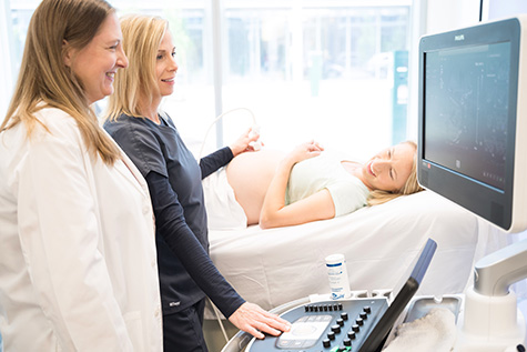 A female doctor standing next to a female ultrasound technician while the technician holds an ultrasound probe against the stomach of a pregnant woman lying down on an exam table as the two providers look at a monitor.
