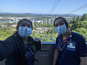 Two women in scrubs and wearing PPE face masks take a selfie at the top of the OHSU tram.