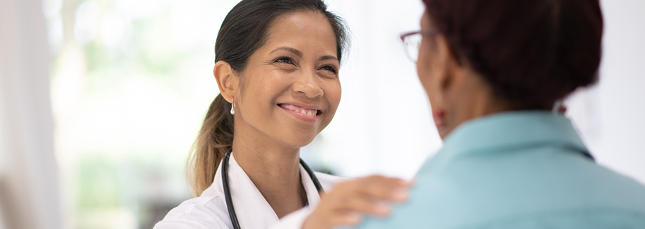 A female doctor smiles while placing her hand on the shoulder of a female patient.