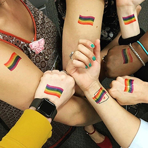 Seven people stretching their arms out displaying matching rainbow flag diversity tattoos.