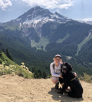 A woman crouching down next to her dog while on a hike with a mountain in the background.