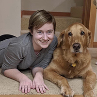 A woman smiling lying on her stomach next to a golden retriever dog.