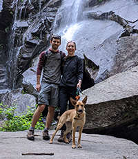 A woman, a man and their dog posing in front of a waterfall while on a hike.