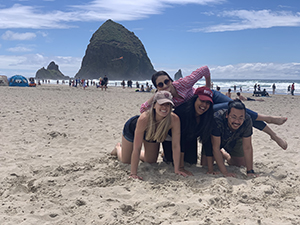 Two girls and one boy crouch down on the beach while a fourth woman lays on top of them, with Haystack Rock in the background.
