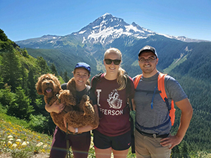A woman holding a dog standing next to another woman and a man (all smiling) while on a hike on a sunny day with Mt. Hood in the background.