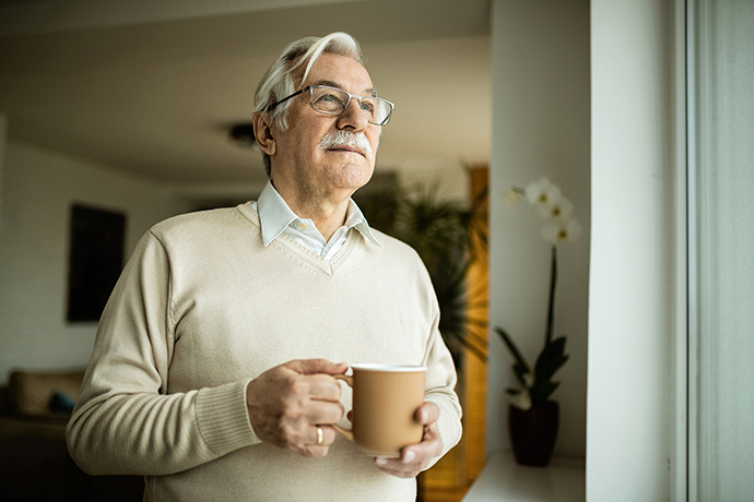 A man with white hair and glasses enjoys a window view while holding a mug.