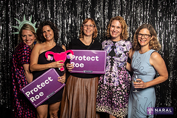 Five women standing next to each other, two of whom are holding up signs that read "Protect Roe."