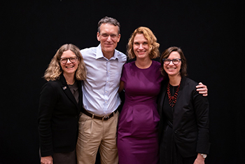 Three women and one man posing with their arms around each others' shoulders.