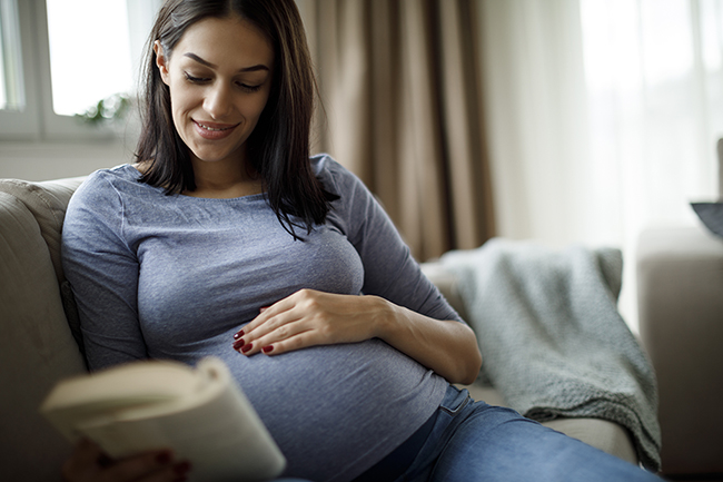 Pregnant woman sits on couch reading a book