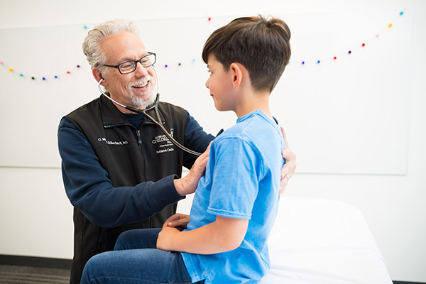 A little boy sits on a hospital exam table as a male doctor uses a stethoscope to listen to his chest.