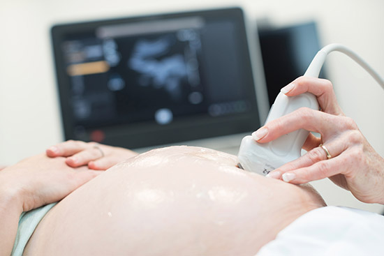 A person's hand holding an ultrasound probe against a pregnant women's belly, with the echocardiogram monitor visible in the background.