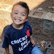 A young boy sitting outside smiling, wearing a t-shirt that says "Chicks Dig Scars."