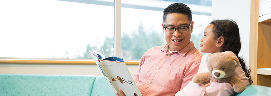 A male health care provider reading a children's book to a little girl in the waiting area of Doernbecher Children's Hospital.