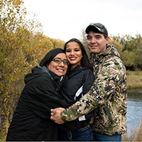 A photo of a teenage girl being hugged by her parents outside by a lake.