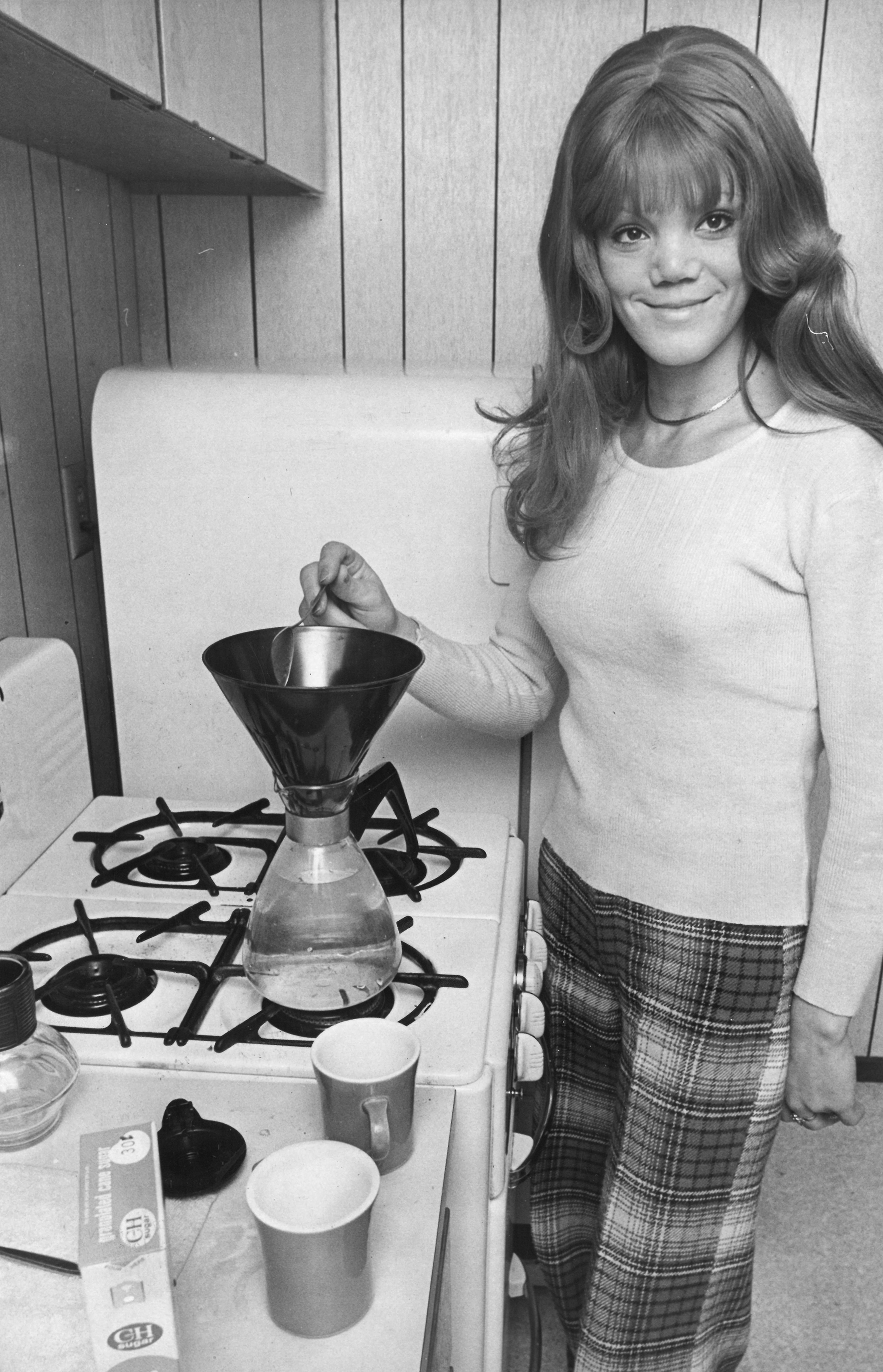 Black and white photograph of Parish standing in a kitchen preparing a chemex with two cups in front of her.