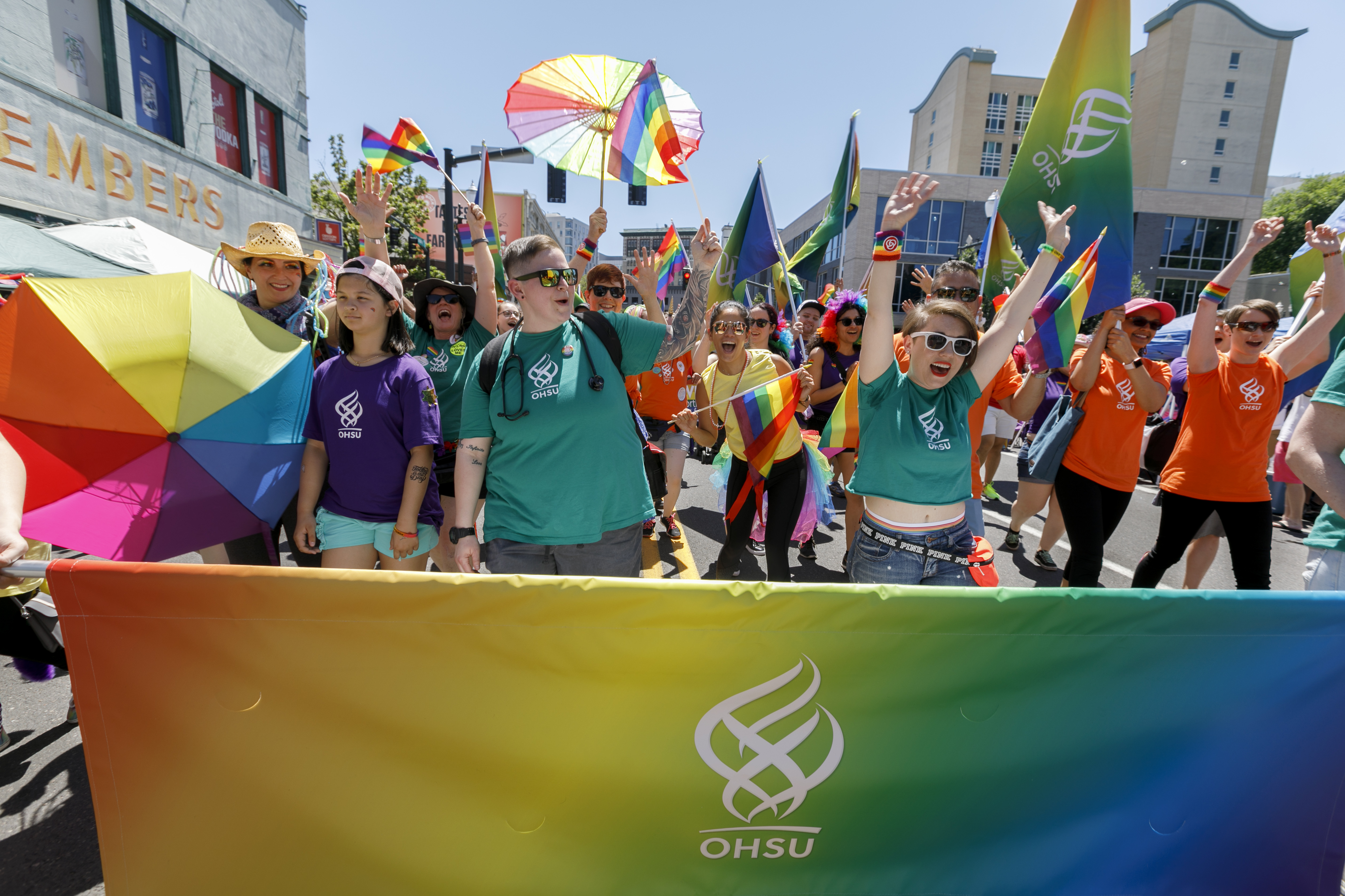 a group of smiling people with different colorful shirts featuring the OHSU logo march behind a rainbow OHSU banner