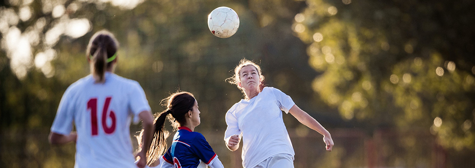 A scene from a soccer game as a teenage girl is about to perform a header, the ball mid-air, while two other girls (one from her team and the other the opposing team) rush towards the ball.
