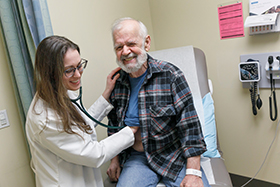 An older man sitting on an exam table smiling while a female doctor listens to his heartbeat using a stethoscope.