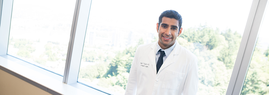Dr. Omar Nazir posing in front of a window in OHSU Hospital, with a view of Portland's South Waterfront behind him.