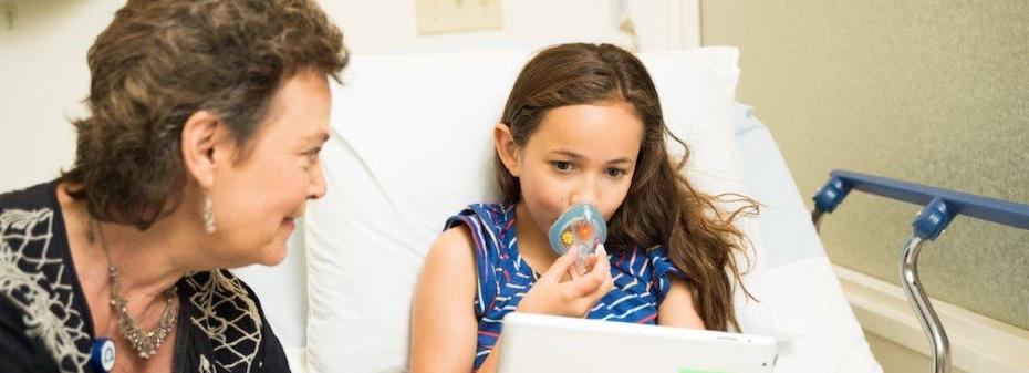 Photo of a health care provider sitting next to a pediatric patient's bed; the child is holding a tablet and has an oxygen mask near their face