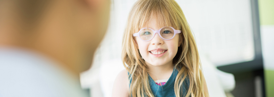 A little girl wearing glasses smiling while her doctor talks to her.