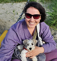 A woman sitting on the beach smiling with a little dog in her lap.