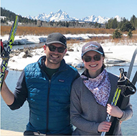 A woman and a man smiling holding skis in front of a lake with mountains in the background.
