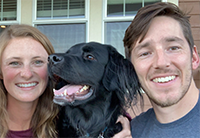A photo of a woman and a man posing with their dog in between them, all seemingly smiling.