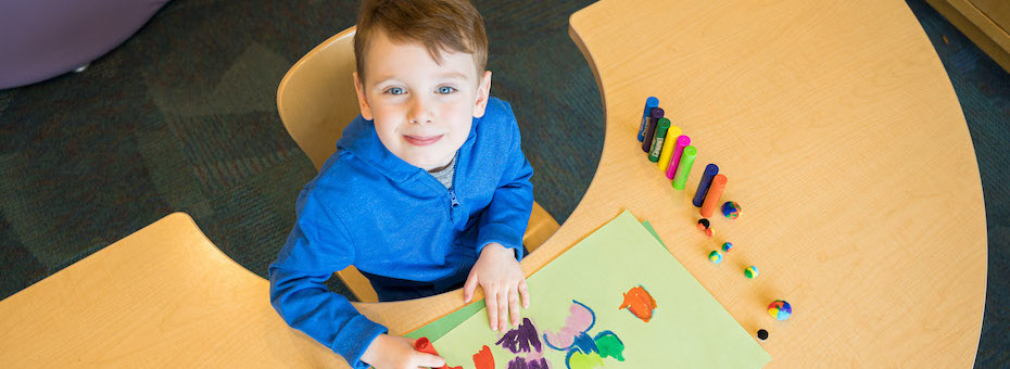 Photo of a child sitting at a curved table coloring on paper with markers