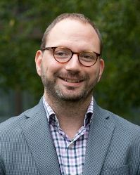 Headshot of a man with glasses, wearing a checkered shirt and a blazer