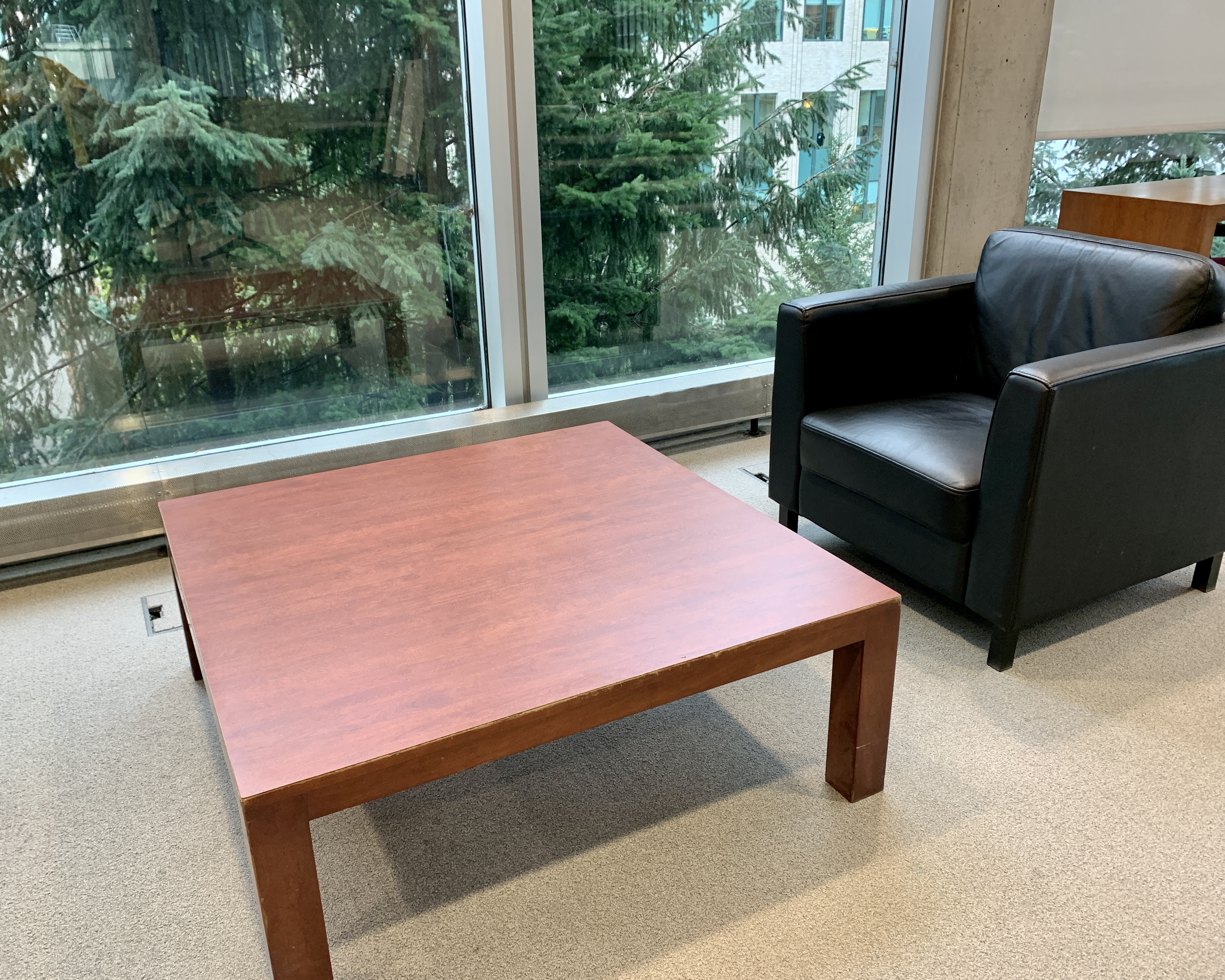 color image of a black leather chair sitting behind a large wooden coffee table. Trees are visible in the background through a large window.