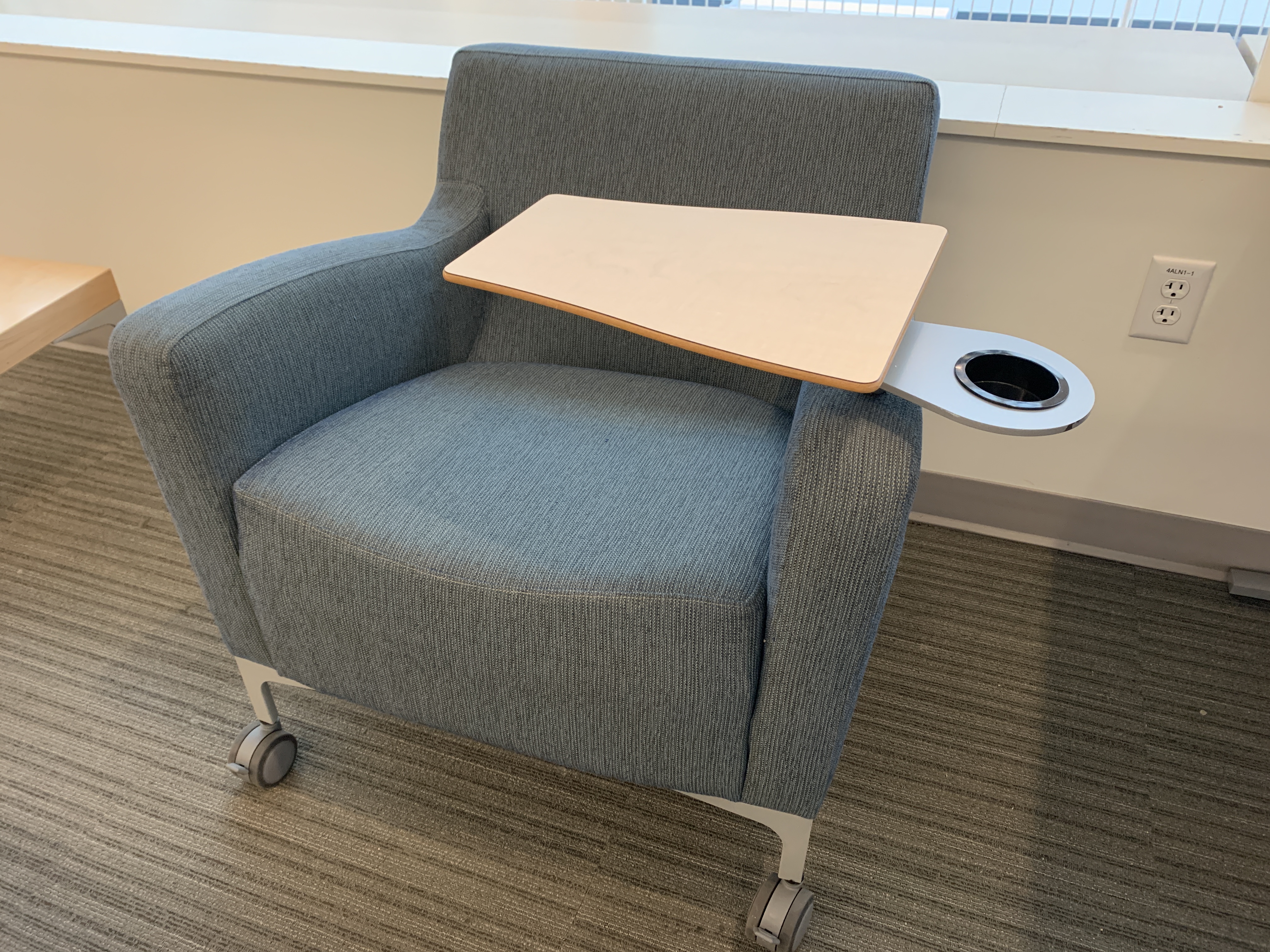 A padded chair with a swivel lap desk and a cup holder sits in a library space.