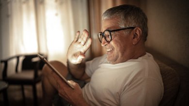 Older man wearing glasses waving to participants on his tablet for a virtual class. 