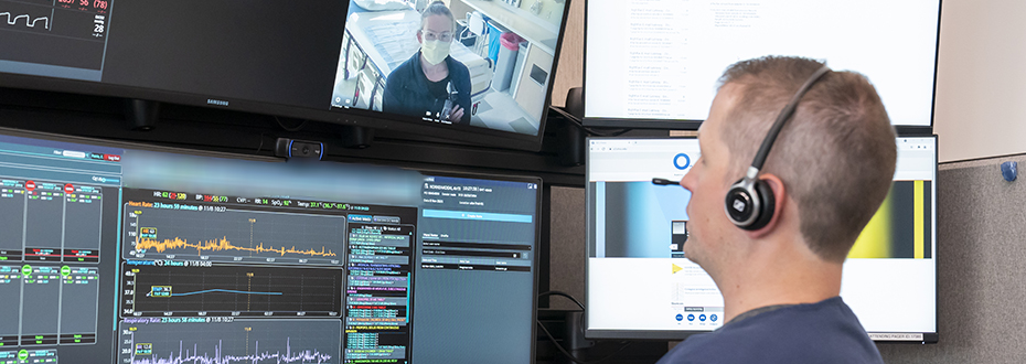 A male doctor sitting at a computer wearing headphones as he videoconferences with a female doctor, visible on his computer screen above a patient's chart notes.