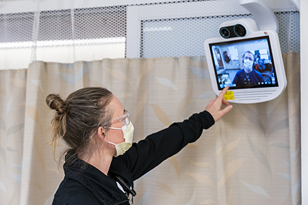 A female nurse touching a monitor screen hanging from the ceiling while she is videoconferencing with another healthcare provider, visible on the screen.