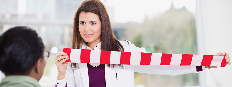 A female doctor sitting in front of a patient and holding a strip of cloth with a pattern of alternating red and white rectangles.