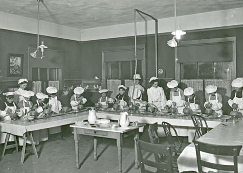 A large room with 11 boys cooking at tables and 4 adult women teaching the class; image is black and white; boys are wearing chef's hats
