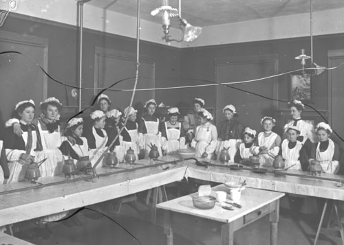 Black and white image showing 13 girls and 4 adult women in a cooking class in a larger room, each student with a burner and pot in front of them and wearing a chef's hat and apron