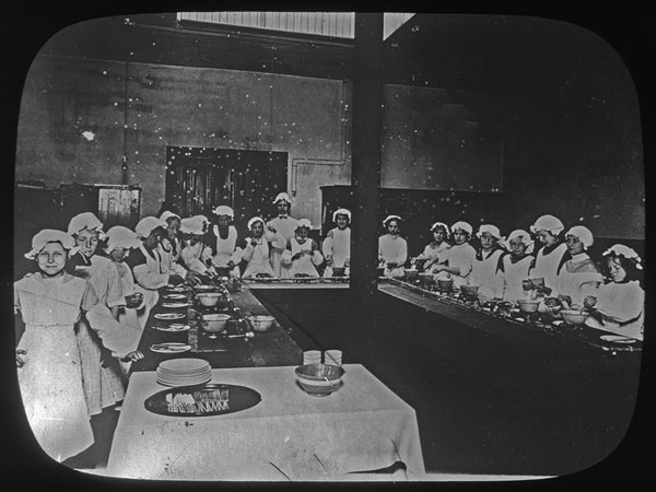 Black and white image showing 19 girls and 1 adult woman in a cooking class, all wearing chef's hats and aprons