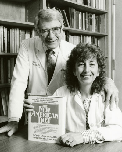 Sonja Connor, seated at a table holding The New American Diet book, William Connor standing behind Sonja with one arm on her shoulder and the other resting on the table; both in white coats in front of a bookshelf