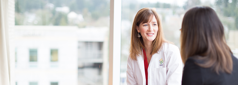 A woman in a white lab coat smiling and speaking with another woman in front of a window.