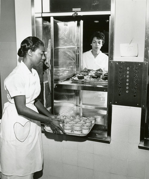 Two young Black women place trays of food into a dumbwaiter