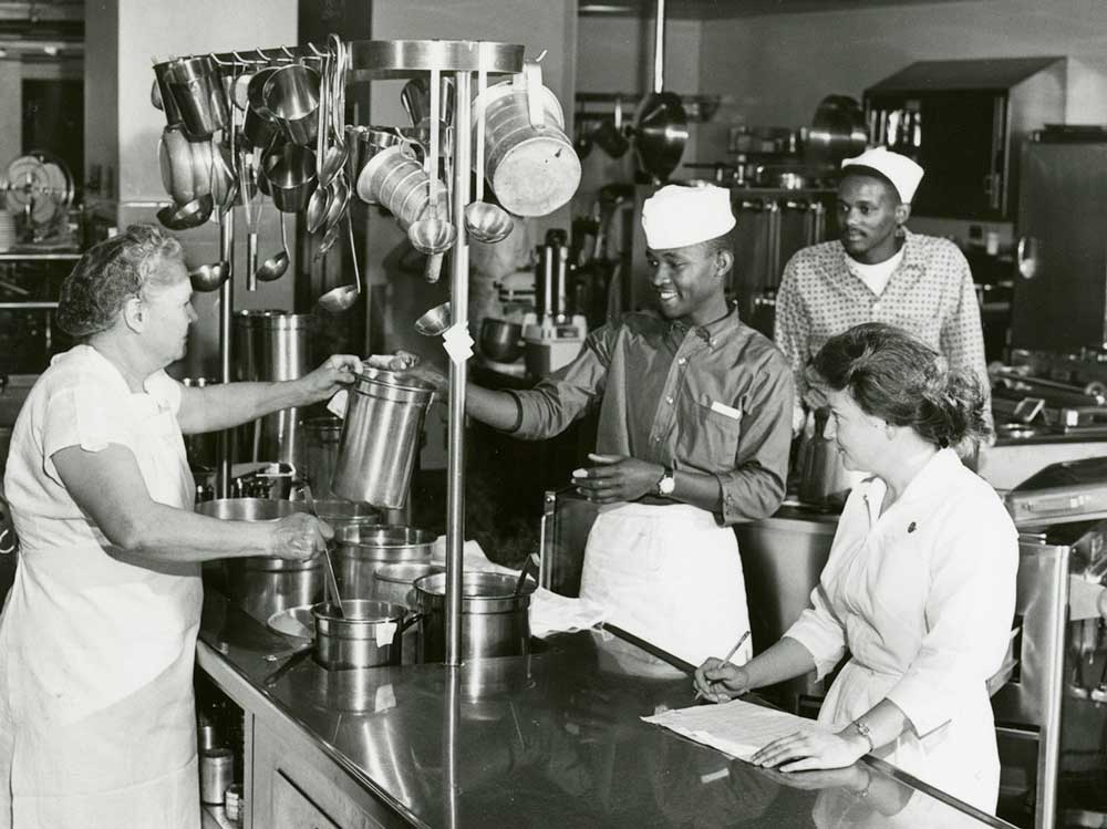 2 young Black men and 2 older White women in the kitchen of Doernbecher Hospital, one woman and handing a pot to one of the men