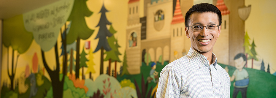 A male doctor smiling while standing in front of a wall mural in the lobby of a children's hospital.