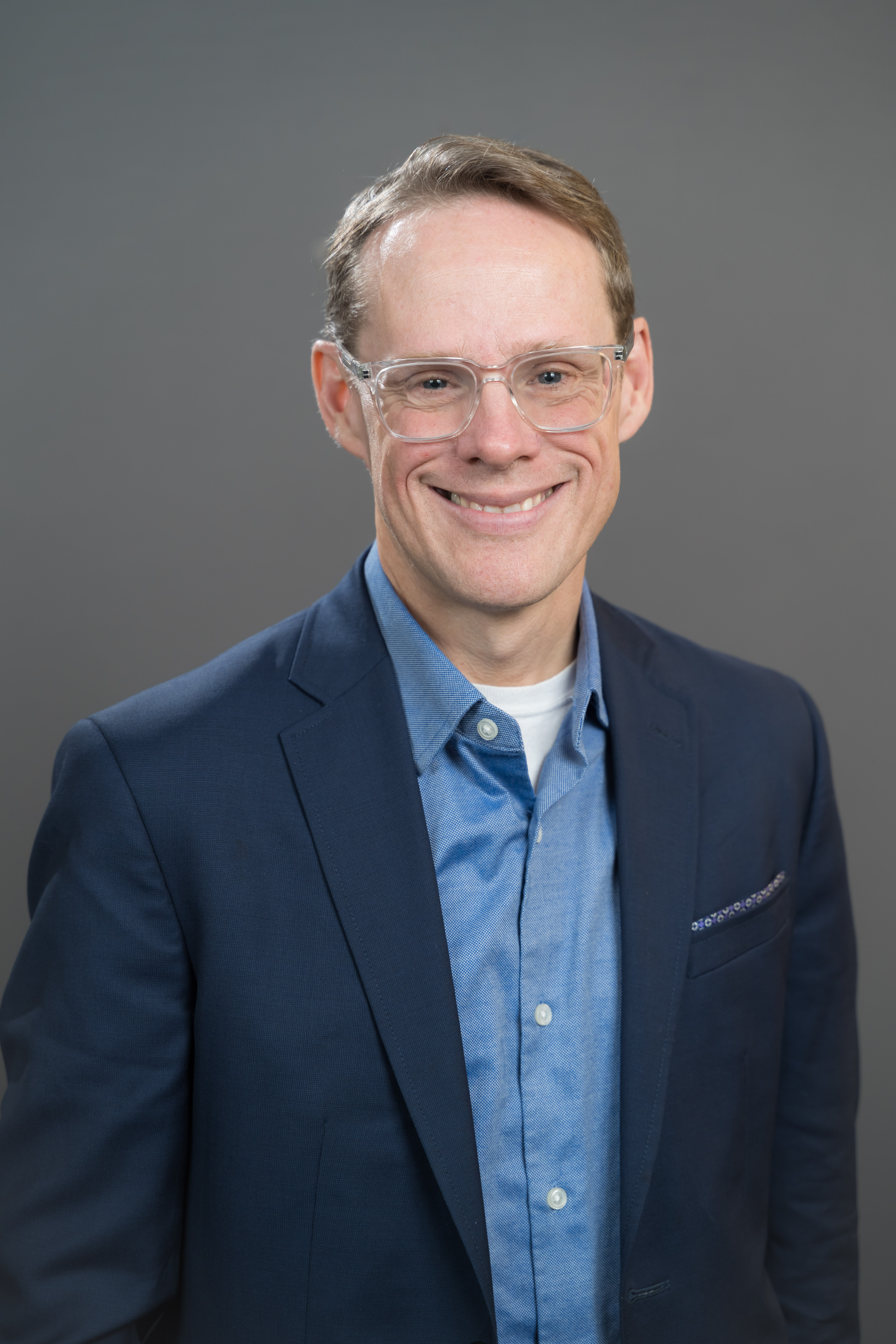 Middle aged man with sandy blonde hair and clear horn-rimmed glasses smiling, wearing a navy blue blazer and light blue button down shirt