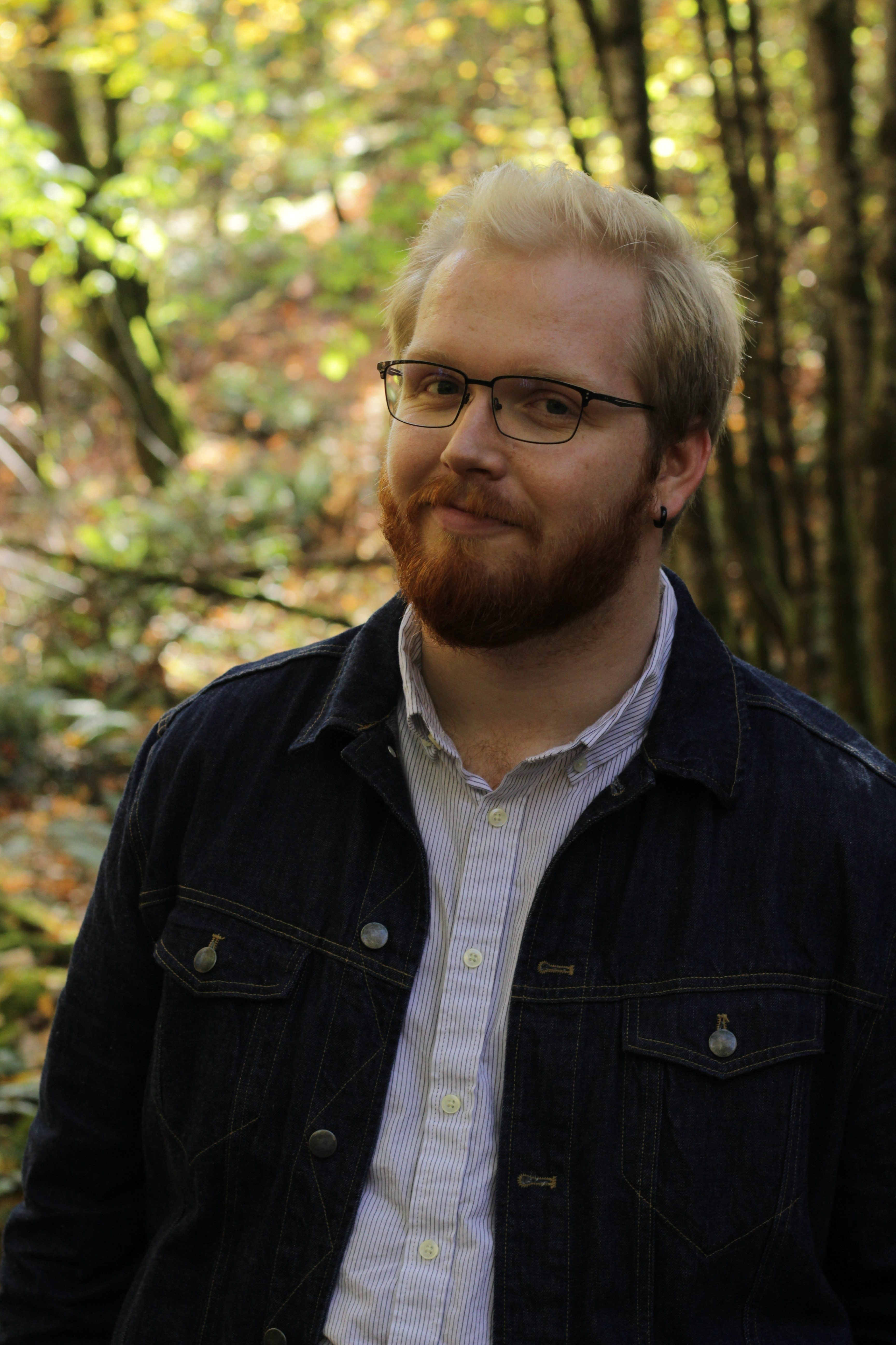 blonde person with glasses and a red beard smirking in a forest pictured from the waist up wearing a dark blue denim jacket and a pinstriped white shirt underneat