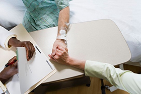 A photo of a hospital patient holding hands with someone else, as a healthcare providers hands are also visible writing something on a medical chart.