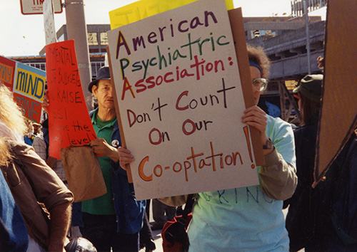 Color photograph of demonstrators. One holds a legible sign that reads "American Psychiatric Association: Don't Count on our Co-optation"